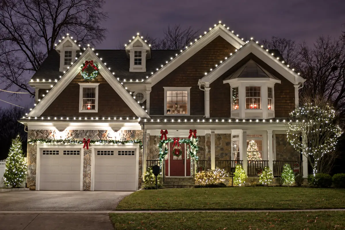 A two-story house at night with white Christmas lights outlining its roof, illuminated wreaths and garlands, and lights covering shrubs and a tree.