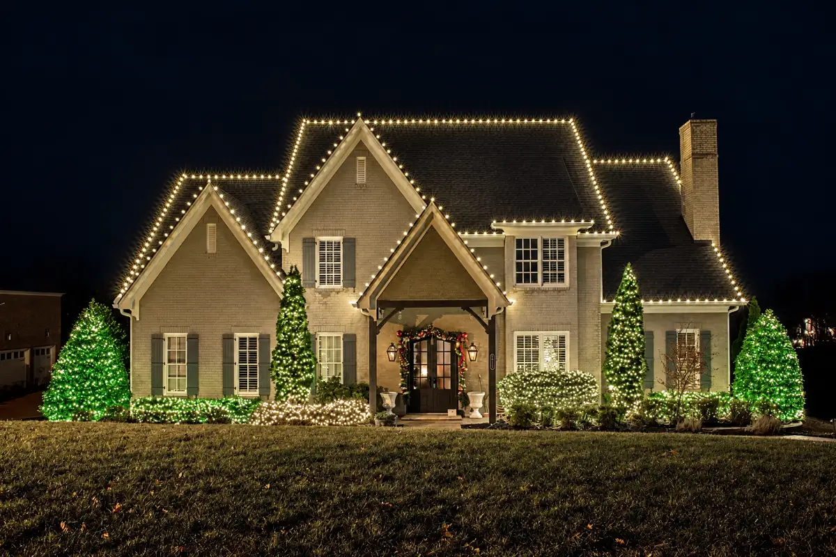 A brick house at night with white holiday lights outlining its roof and green and white lights covering front-yard trees and shrubs.